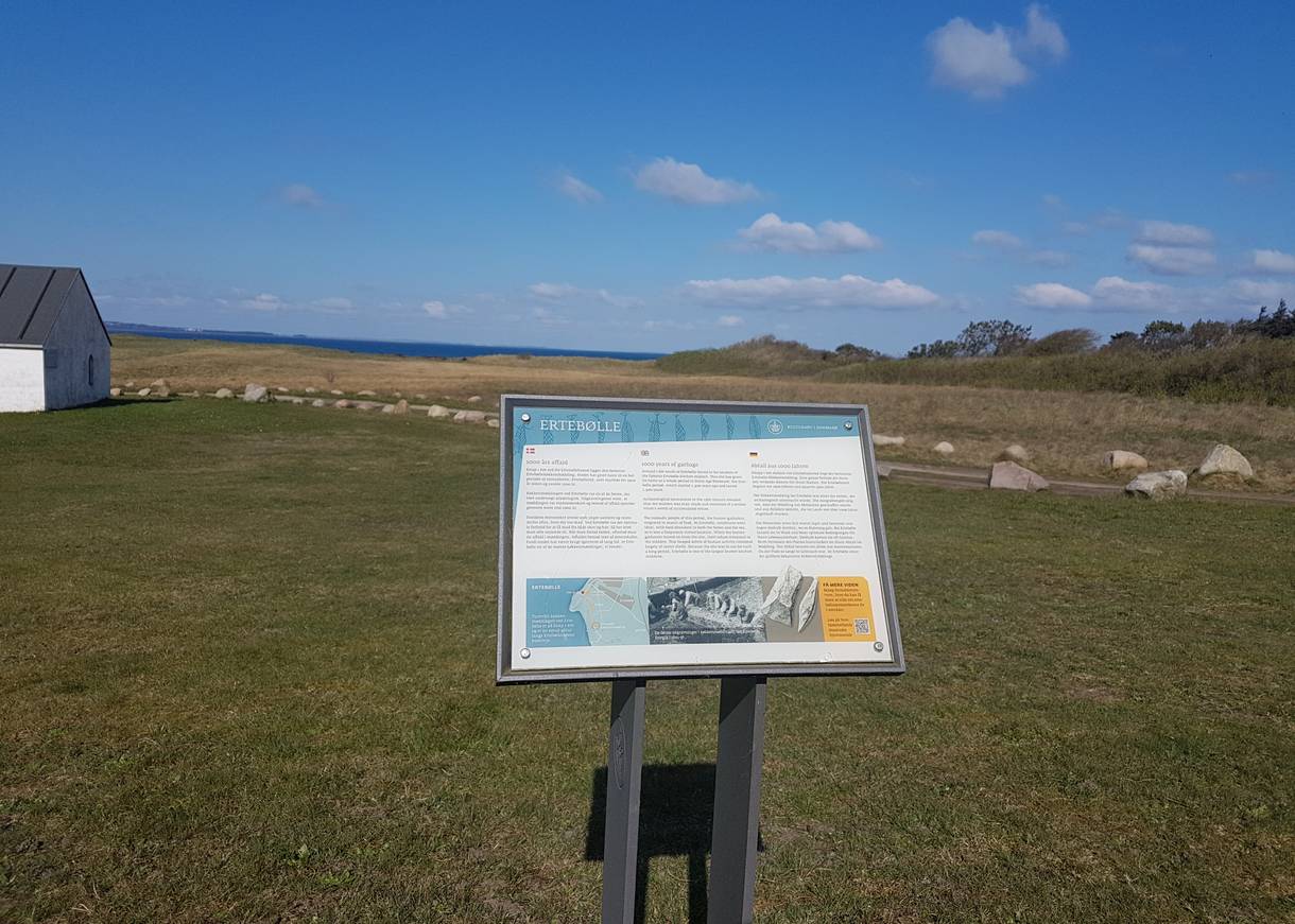 Info-Schild vor einer Wiese, am Bildrand ein kleines, weißes Haus