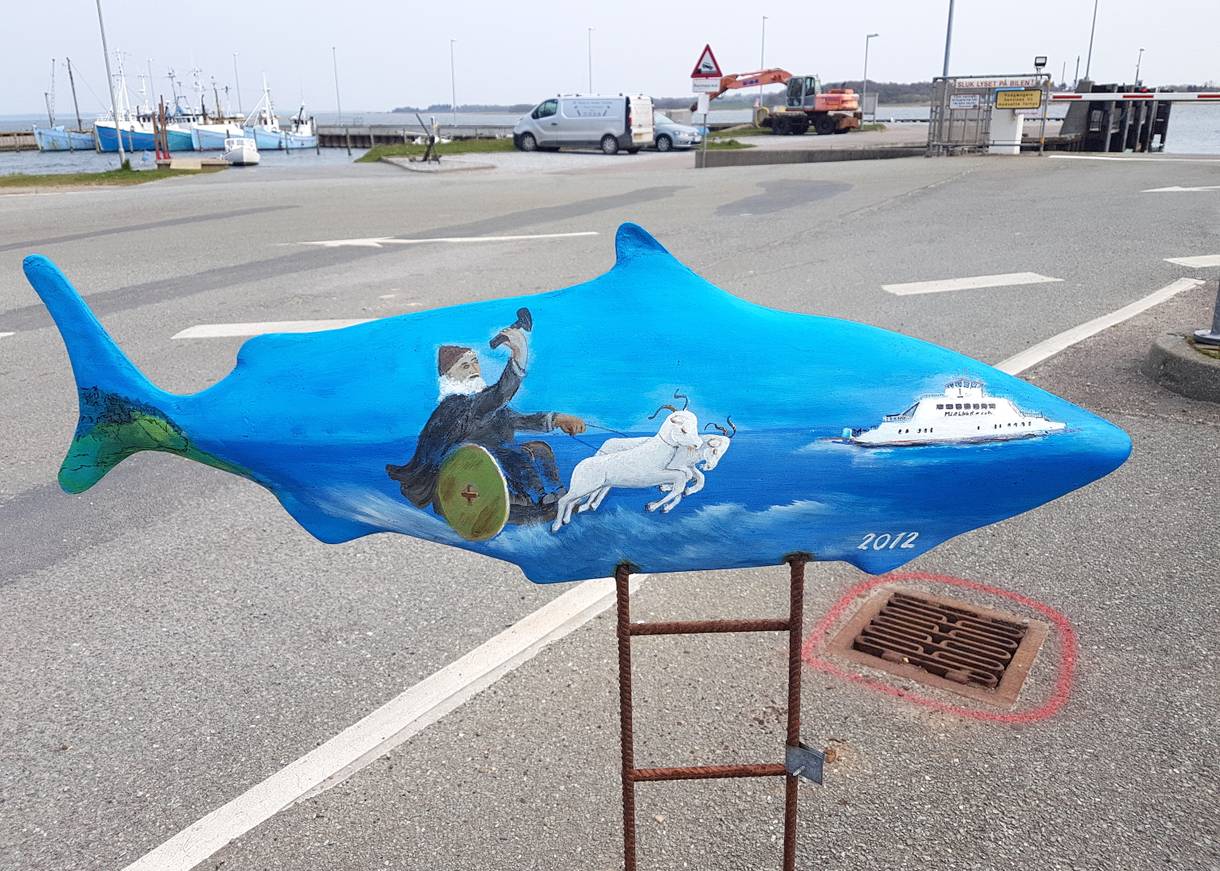 Blaue Fischskulptur auf einem Metallständer, dahinter ein Parkplatz am Hafen