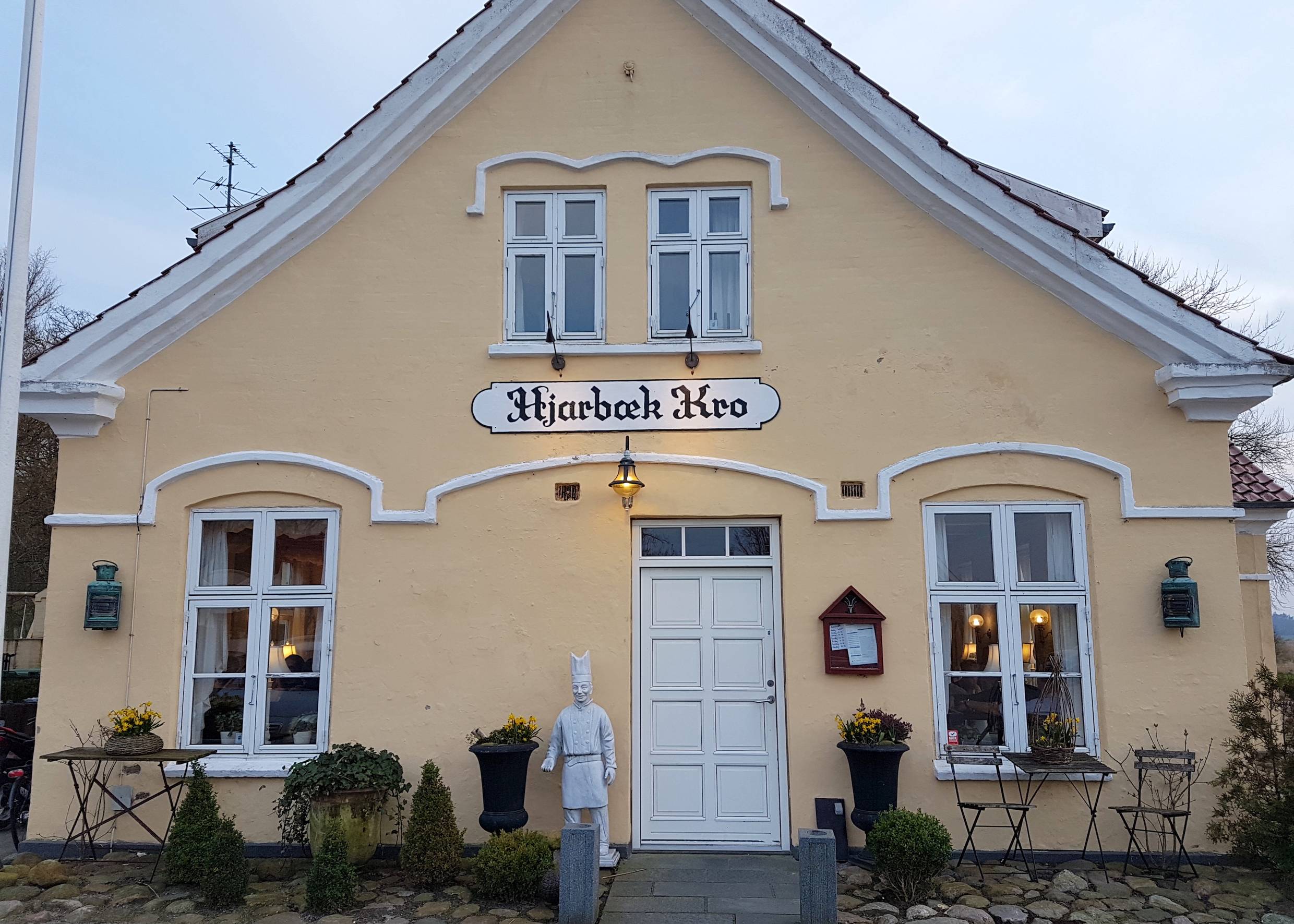 Hellgelbes Haus mit weißen Fenstern und Türen, über der Tür ein geschwungenes Namensschild 