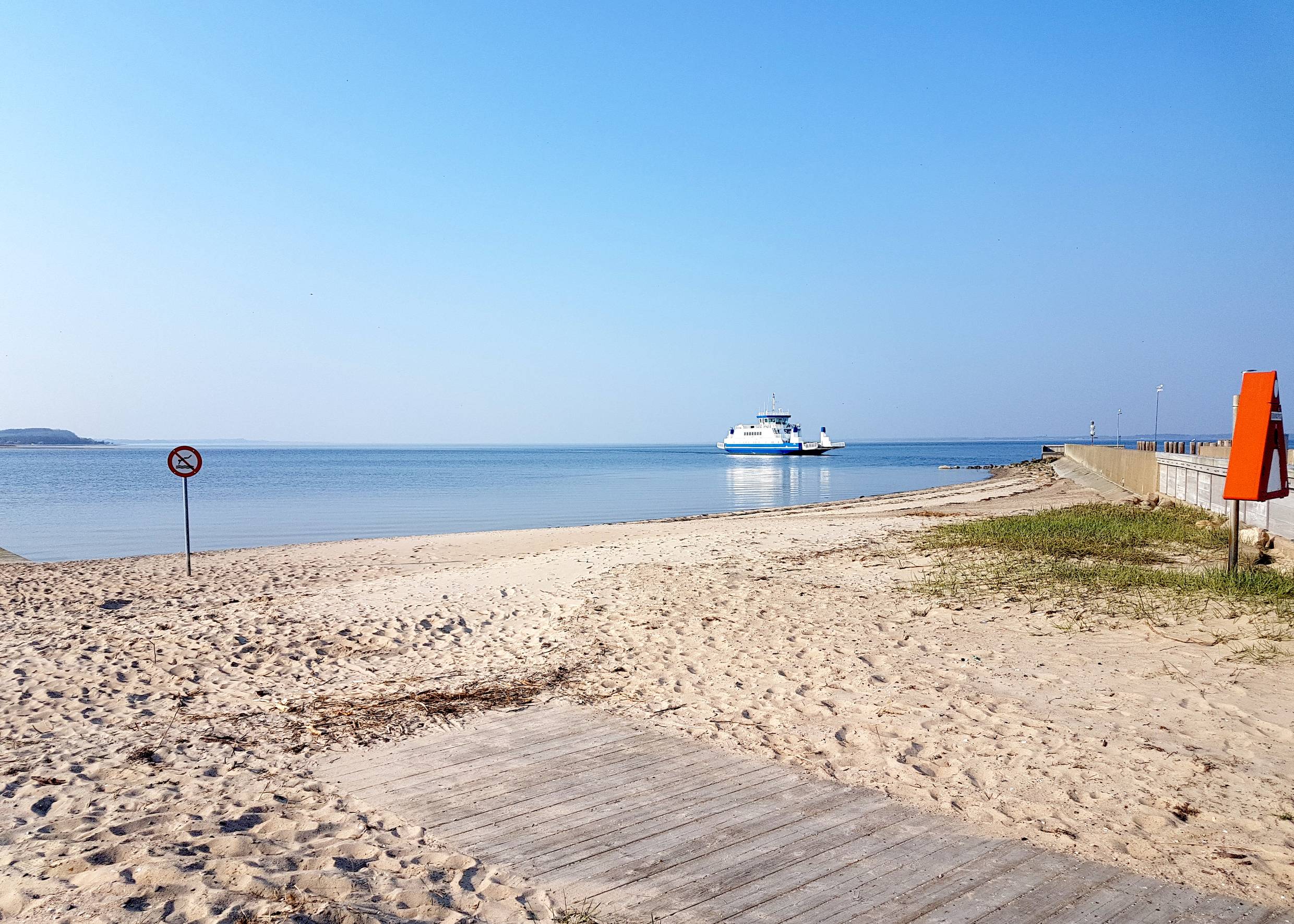 Sandstrand mit barrierefreiem Zugang, dahinter eine kleine Fähre auf dem windstillen Fjord