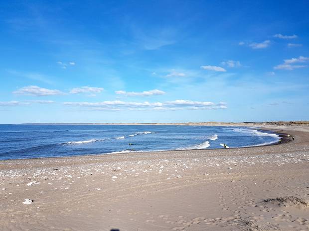 Eine Strandbucht mit Wellen, im Vordergrund eine Person mit SUrfbrett unter dem Arm