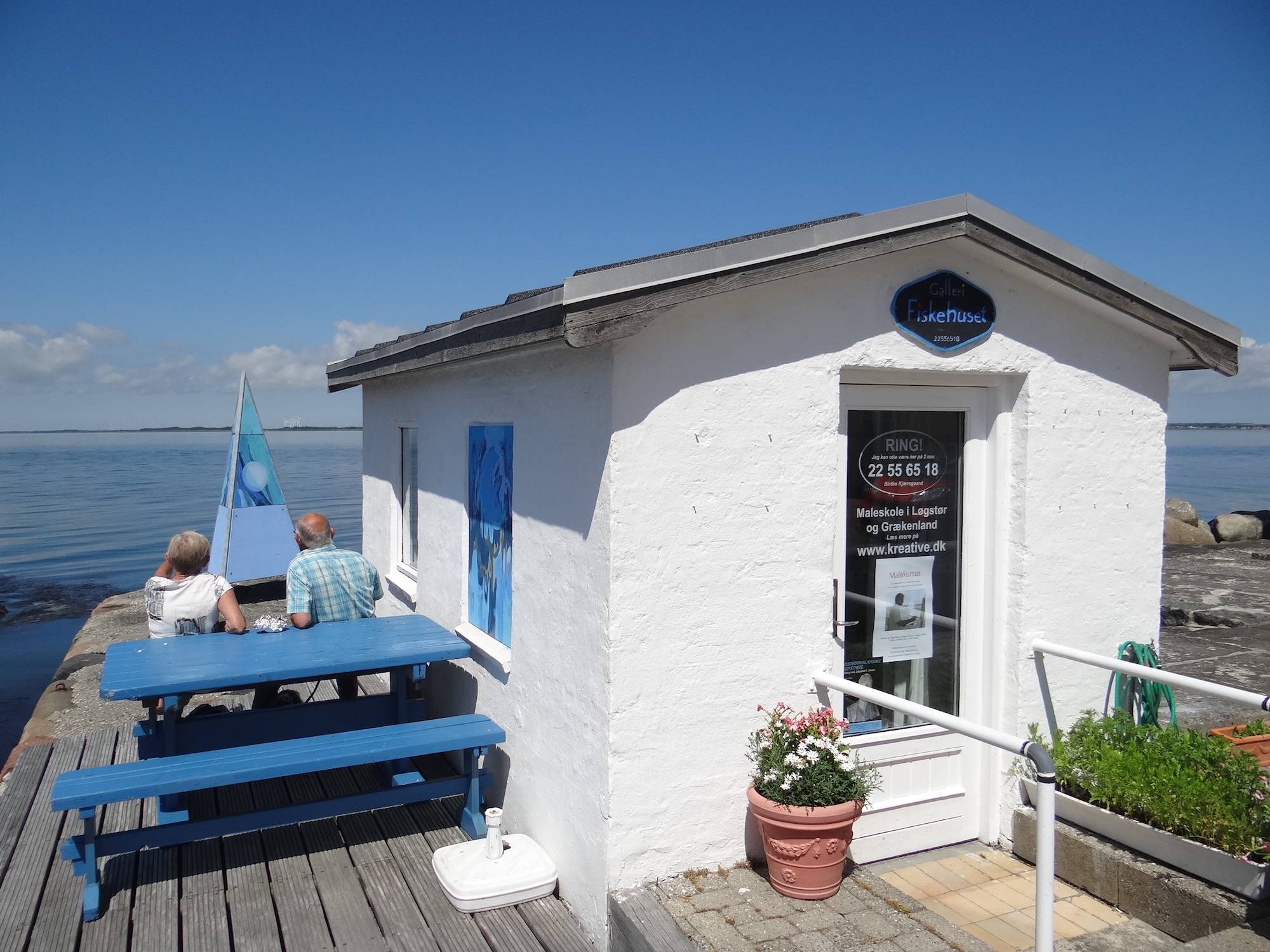 Weiße Steinhütte mit blauen Picknickbänken auf einer Holzterrasse am Wasser