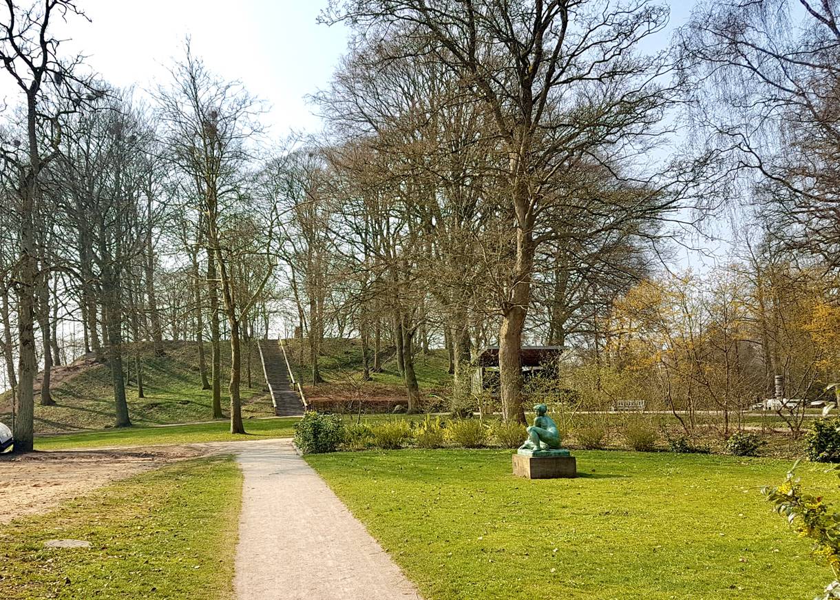 Blick auf einen Wall in einem Park, frühlingsgrüne Bäume und Skulpturen am Weg
