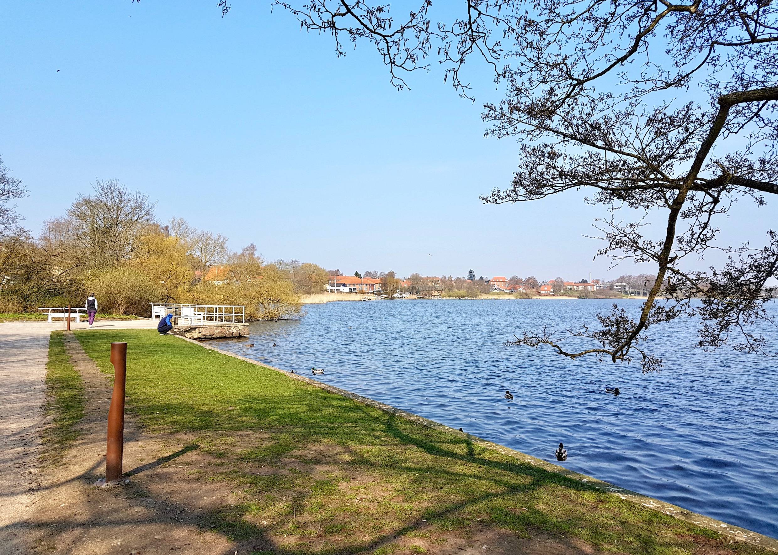 See mit Enten und einem kleinen Anleger, im Hintergrund Häuser am Ufer