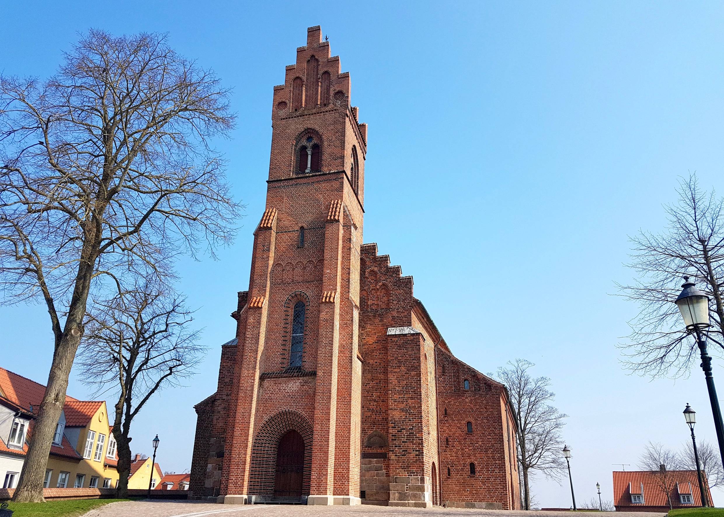 Große rote Backsteinkirche mit eckigem Turm über dem Portal, dahinter blauer Himmel