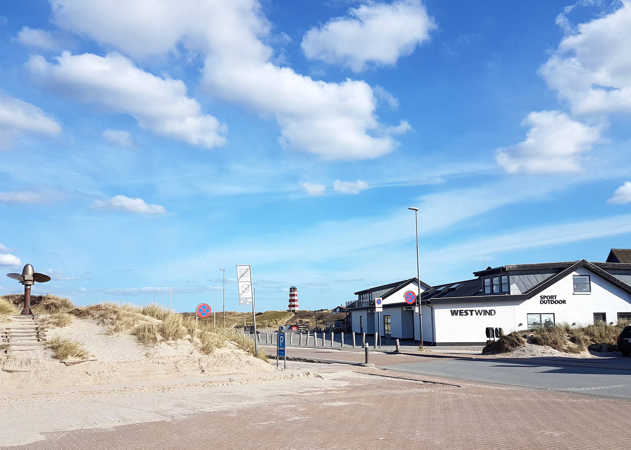 Blick vom Parkpltz auf ein Sportgeschäft, dahinter eine Art Leuchtturm auf einer Düne