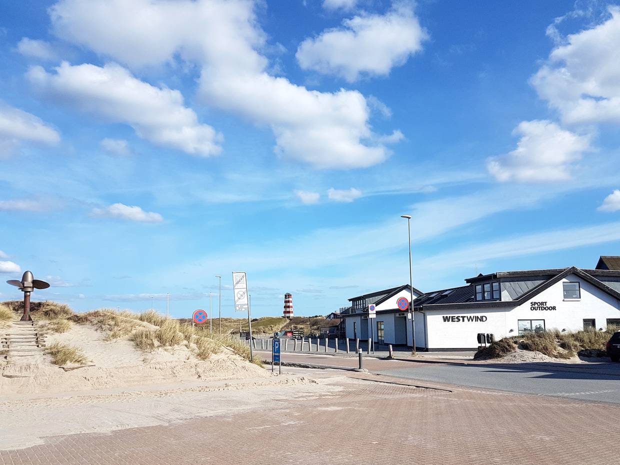 Blick vom Parkpltz auf ein Sportgeschäft, dahinter eine Art Leuchtturm auf einer Düne