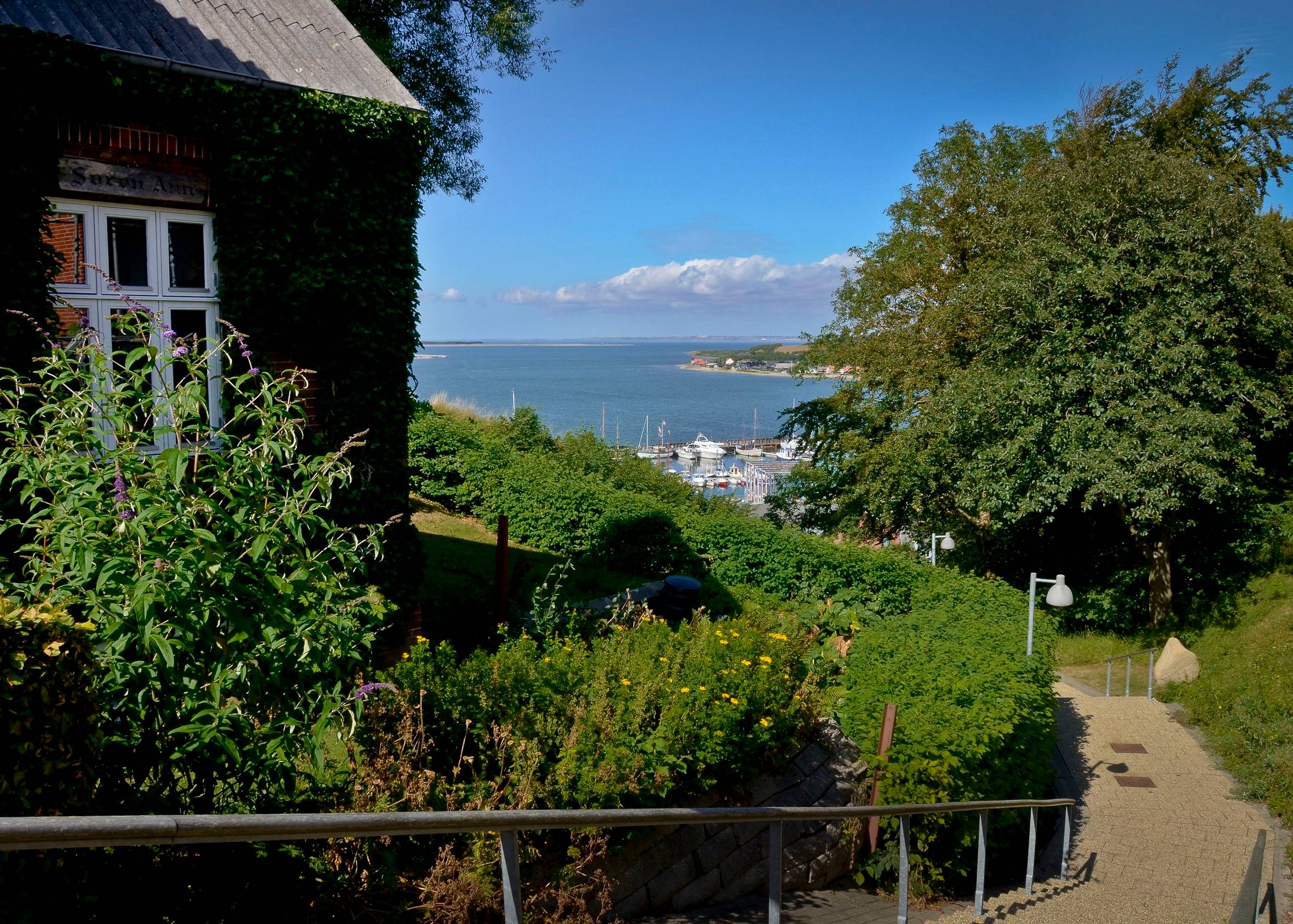 Fußweg zwischen Häusern und Gärten mit Blick auf einen Hafen mit Segelbooten