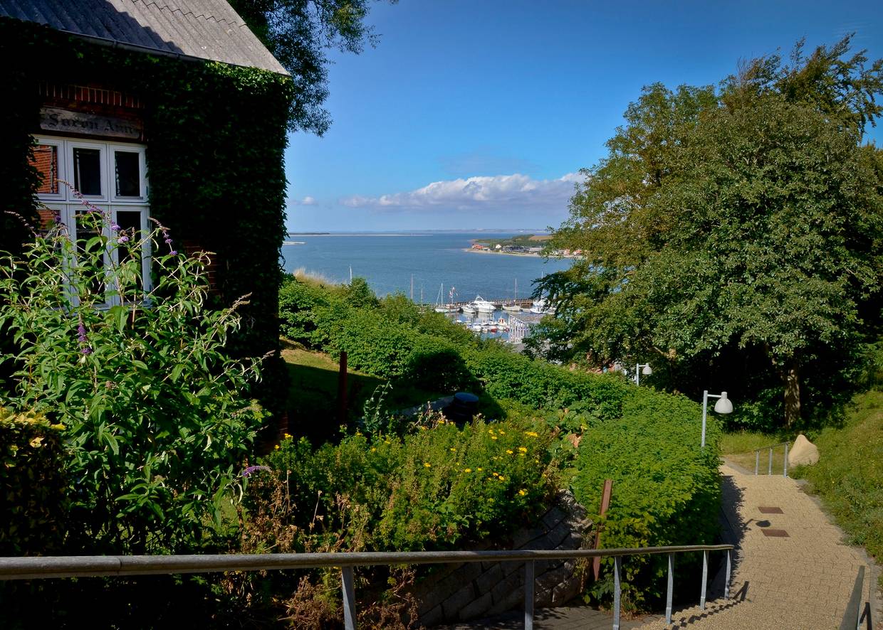 Fußweg zwischen Häusern und Gärten mit Blick auf einen Hafen mit Segelbooten