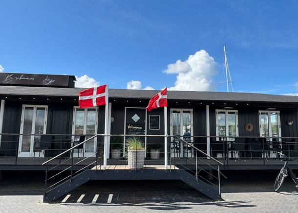 Ein Restaurant in einem schwarzen Holzgebäude mit zwei Fahnenmasten davor und Sitzplätze