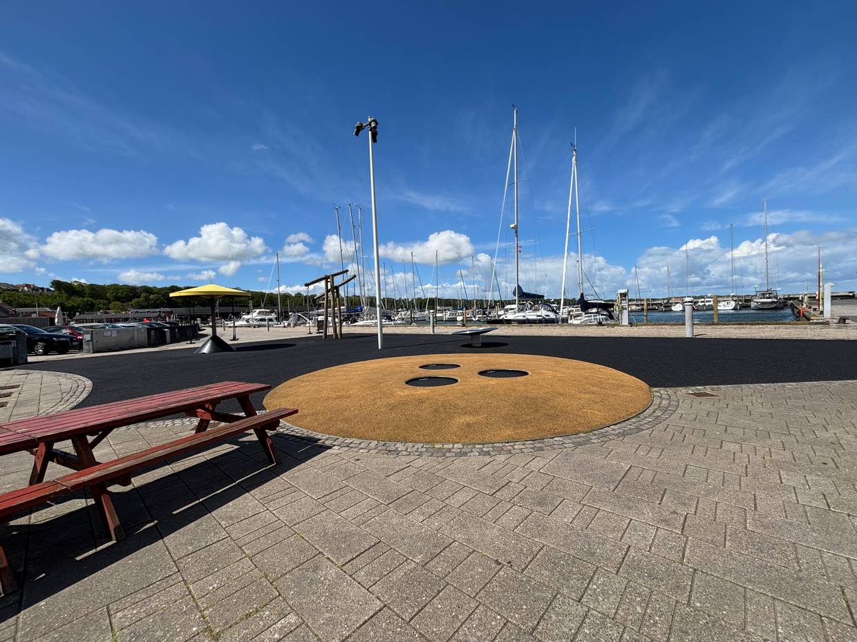 Blick über einen Hafen, im Vordergrund einen Trampolin, ein Picknicktisch und weitere Spielgeräte