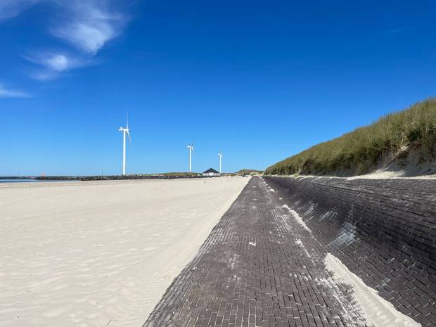 Blick auf eine Strandlandschaft mit weißem Sand, drei große Windmühlen und einen Weg aus Ziegelsteinen an der Düne entlang