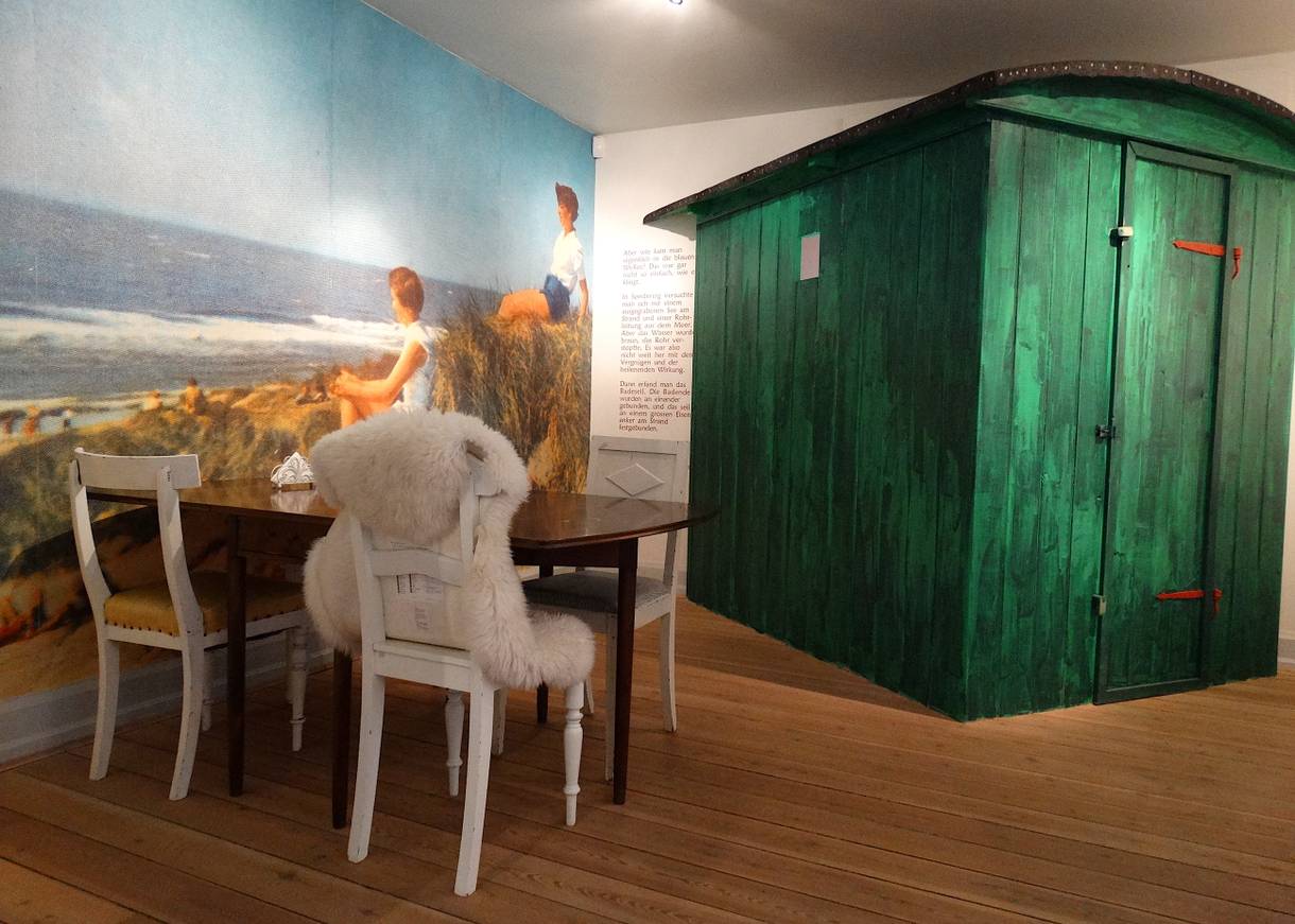 Esttisch, dahnter eine grüne Badehütte, an der Wand Fototapete mit Dünen