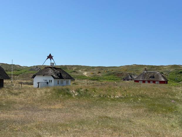 Reetgedeckte Ferienhäuser in einer Dünenlandschaft, dahinter eine Bake
