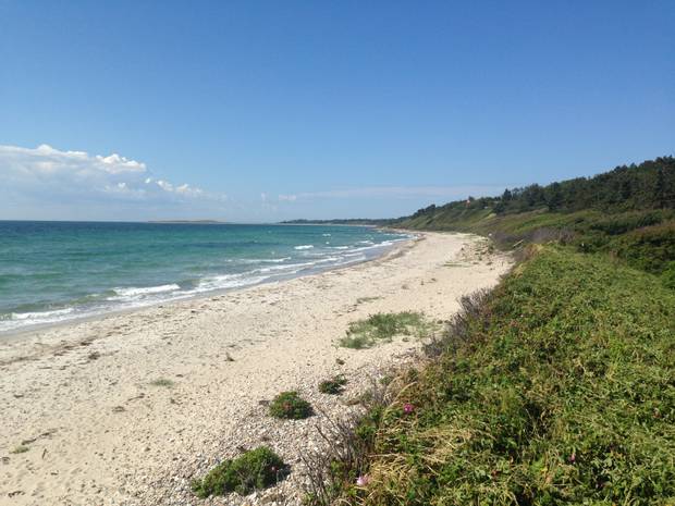 Eine Langgestreckte Bucht mit einem schmalen Sandstrand, dahinter grüne Hügeln