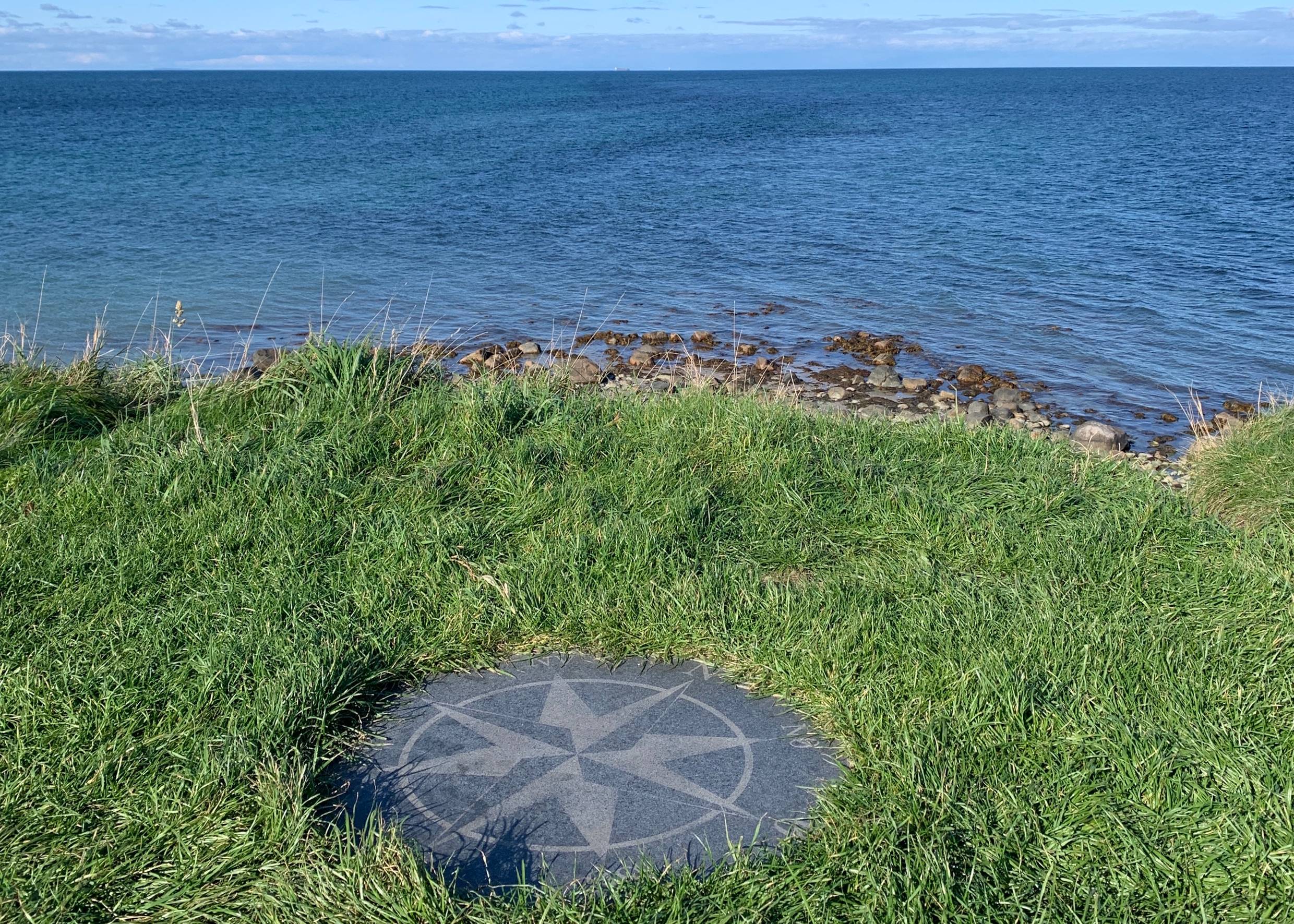 Kompassrose im Gras vor der steinigen Küste