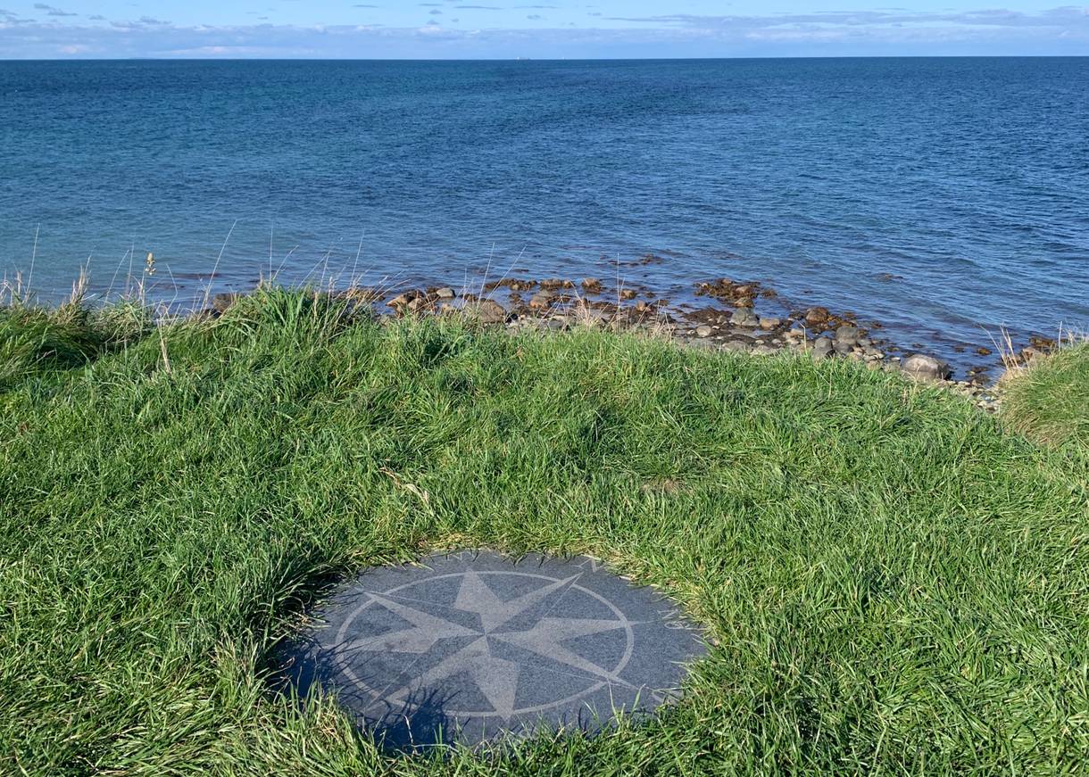 Kompassrose im Gras vor der steinigen Küste