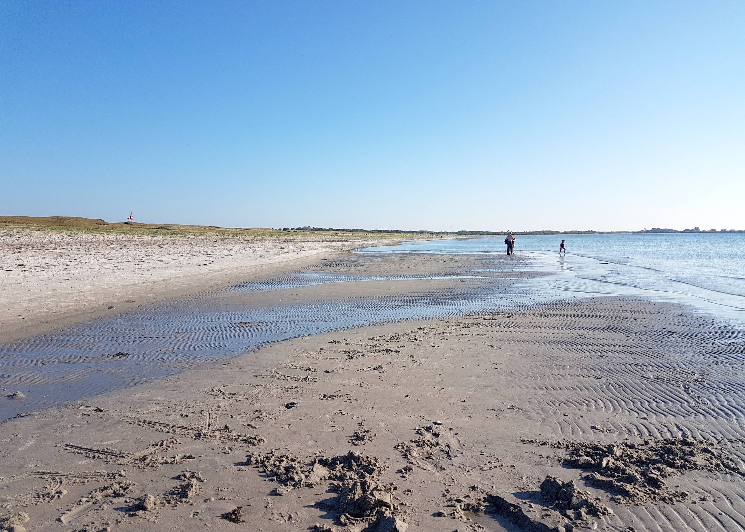 Flacher Sandstrand mit einer Sandbank und Spaziergängern im Hintergrund